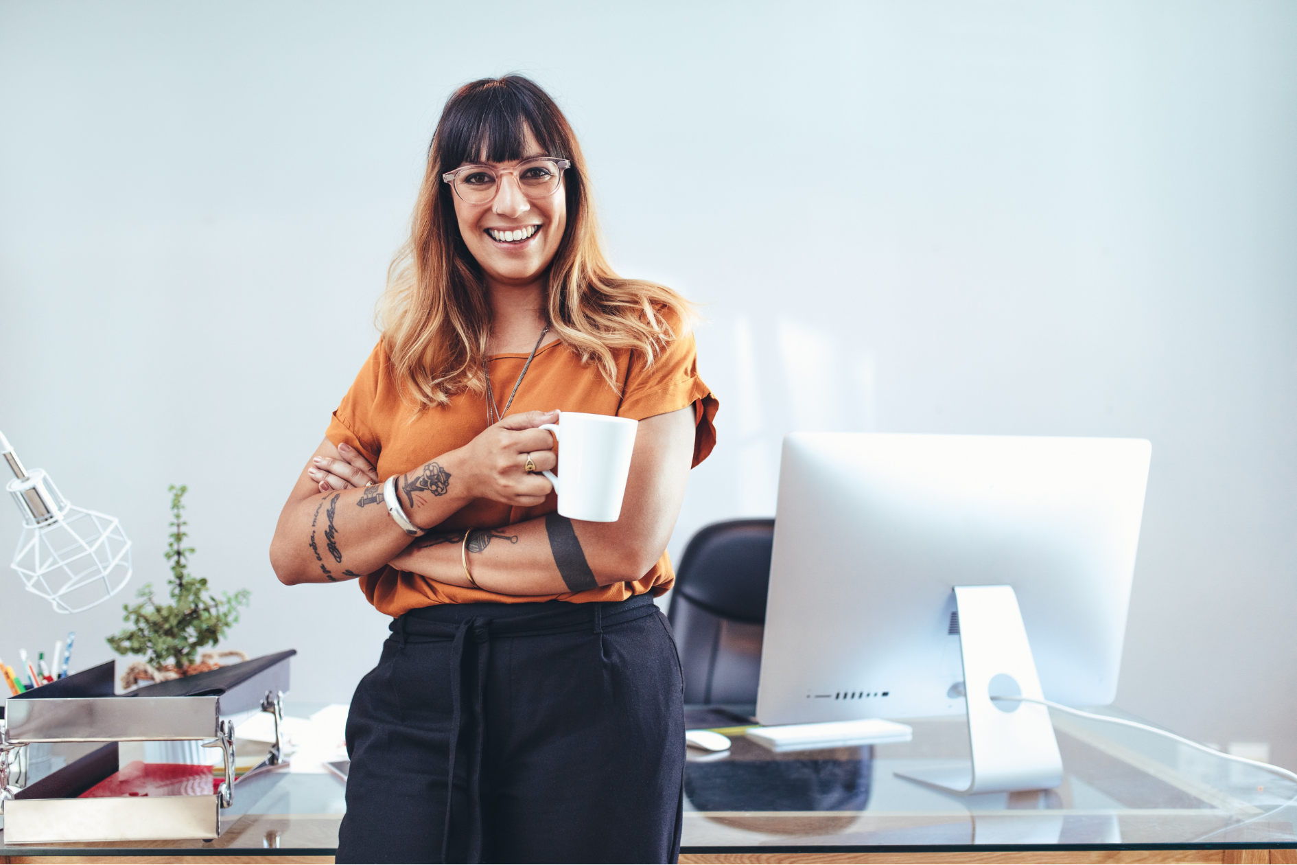 image of female creative entrepreneuer standing and leaning against desk with apple computer and holding a coffee mug